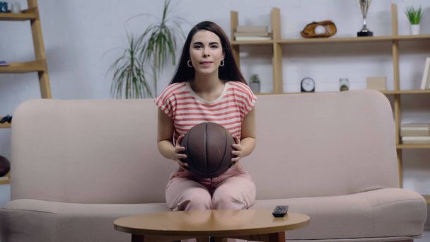 mujer fan del deporte positivo y concentrado viendo partido de baloncesto en la televisión mientras está sentado en el sofá con pelota - Foto, Imagen