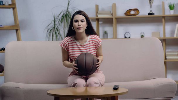 focused basketball fan watching game on home tv while sitting on couch with ball - Photo, Image