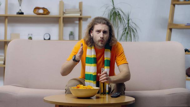 worried man in striped scarf pointing with hand while watching sport competition near beer and chips - Photo, Image