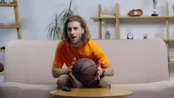 захоплений спортивний фанат з м'ячем сидить на дивані вдома і дивиться баскетбольний матч по телевізору
 - Фото, зображення