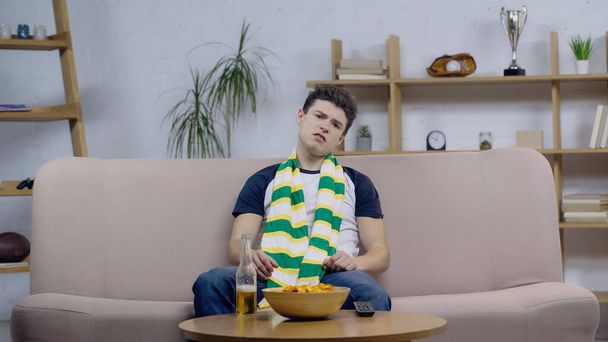 frustrated sport fan sitting on couch near beer and chips while watching game on tv - Foto, afbeelding