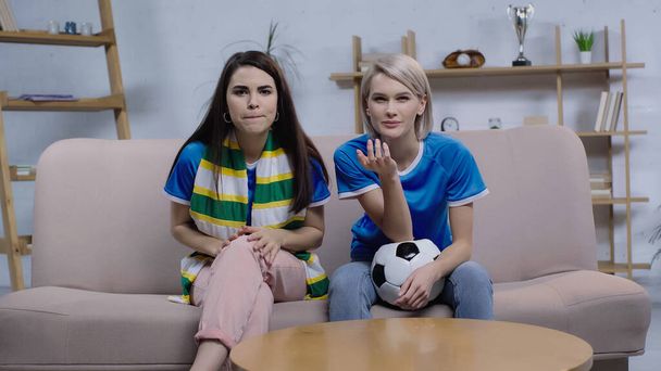 напружені жінки сидять на дивані з футбольним м'ячем і смугастим шарфом і дивитися футбольний матч на телебаченні
 - Фото, зображення
