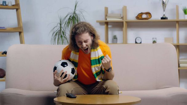 fanático del fútbol lleno de alegría gritando y mostrando gesto de victoria mientras está sentado en el sofá con pelota de fútbol - Foto, Imagen