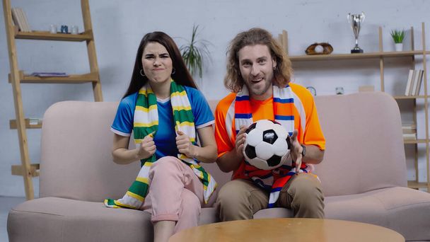 worried woman in striped scarf holding clenched fists for luck near excited friend with soccer ball - Foto, Imagem