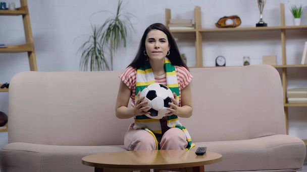 εστιασμένη γυναίκα με ριγέ φουλάρι κρατώντας μπάλα, ενώ βλέποντας ποδόσφαιρο αγώνα στην τηλεόραση στο σπίτι - Φωτογραφία, εικόνα
