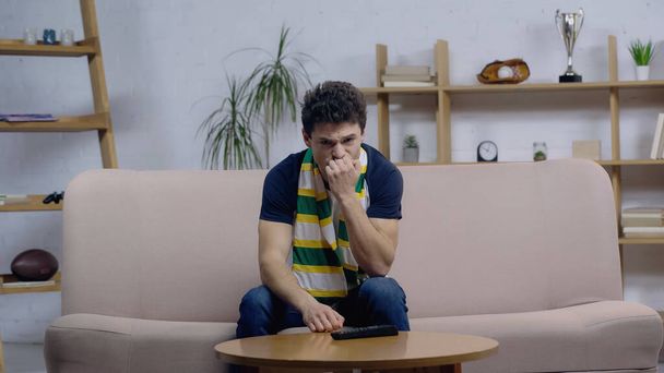 frustrated sport fan in striped scarf sitting on sofa at home and watching match on tv - Foto, Imagem