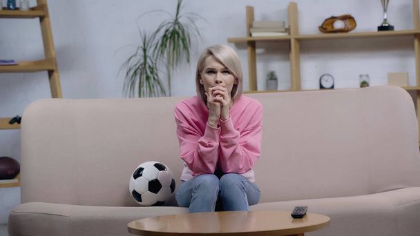 nervioso deporte fan mujer cogido de la mano cerca de la cara mientras mira partido en sofá cerca de fútbol pelota - Foto, Imagen
