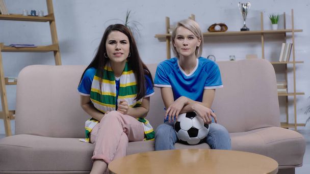 gespannen en geconcentreerde vrouwen zitten op de bank met voetbal en kijken naar wedstrijd op tv - Foto, afbeelding