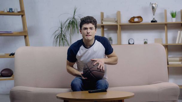 tense sport fan sitting on couch with ball while watching basketball game on home tv - Photo, Image