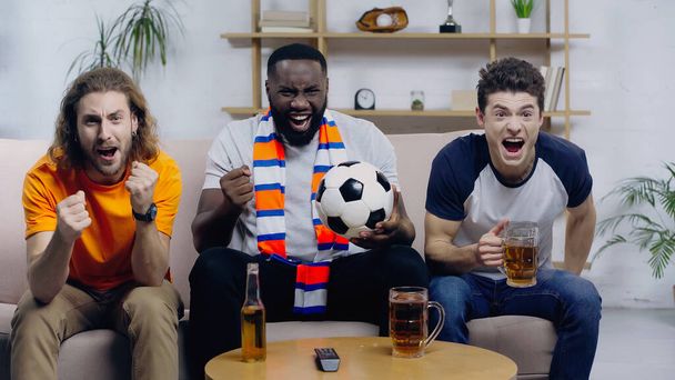 african american man screaming and showing win gesture while watching football match with excited friends - Photo, Image