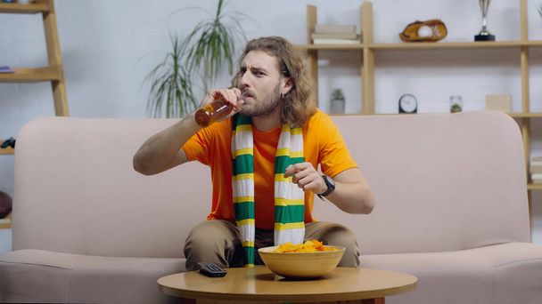 deporte ventilador en la bufanda a rayas beber cerveza mientras ve el juego en la televisión en casa - Foto, Imagen