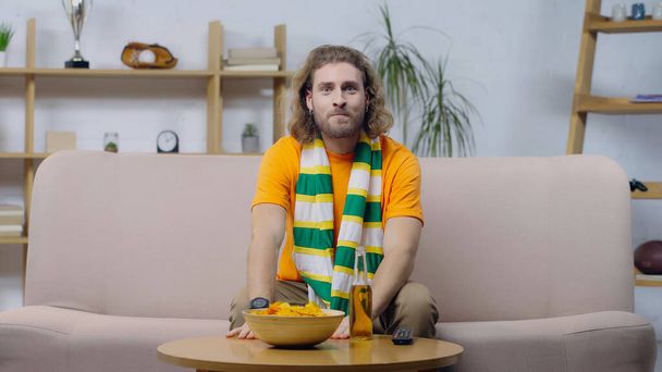 anxious man in striped scarf watching sport game on tv near chips and beer on coffee table - Photo, Image