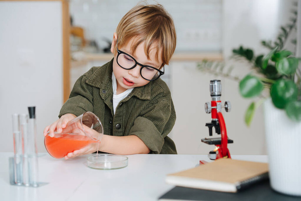 Cute little boy doing science project, filling a glass chemical dish from a beaker. Behind a table with books and notepads. - Foto, imagen