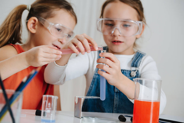 Happy little girls doing science project, they are mixing two colored liquids, pouring from one flask into another. Both wearing protective glasses. - Photo, image