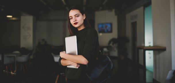 Young attractive female student in casual clothes with backpack holding netbook while standing in modern workspace and looking at camera - 写真・画像