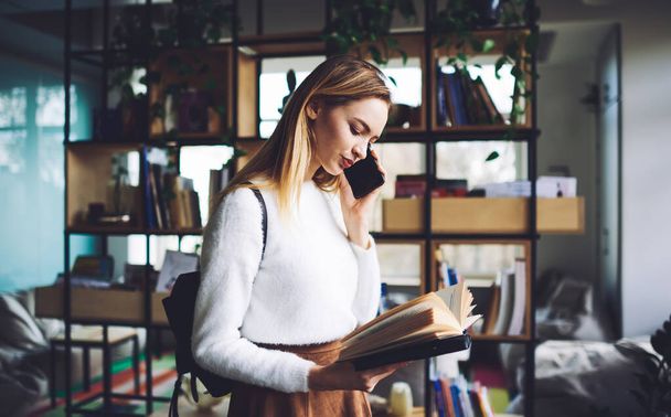 Side view of young lady standing in modern bookstore with opened book in hand while talking on phone and wearing casual outfit with backpack in daytime - Фото, зображення
