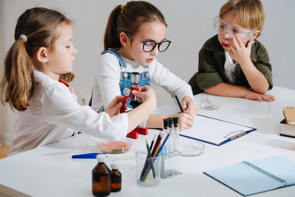 Tired little kids doing home science project, girl is documenting the process. All wearing glasses, sitting behind the table. Chemical stuff on the table. - Foto, Imagen