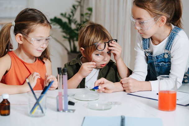 Amusing kids doing home science project, looking at leaf in tweezers. Funny boy looking through magnifying glass. All behid table, wearing glasses. Chemical glassware and colored liquids on the table. - Фото, изображение