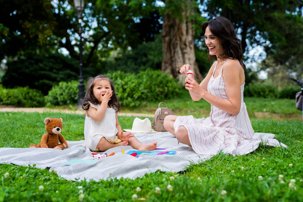 Cheerful female with toothy smile in striped casual dress with bubble blower having picnic with baby girl with teddy bear and crayons in park - Fotó, kép