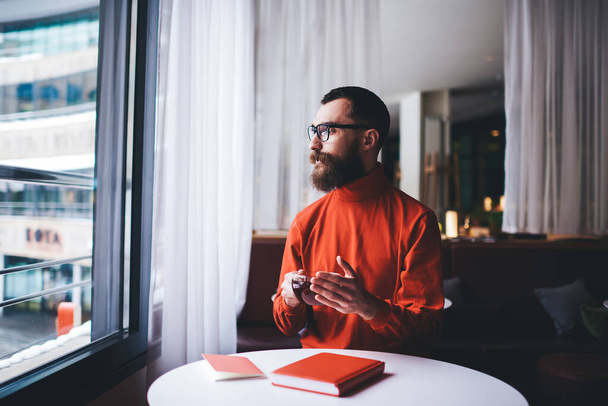 Serious bearded male in casual clothes sitting at round table with cup of hot tea looking out window spending free time alone - Foto, Bild
