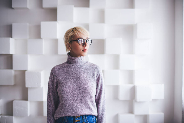 Young calm female in casual outfit with eyeglasses and short blond hair looking away standing on white background with cubes - Foto, Imagen