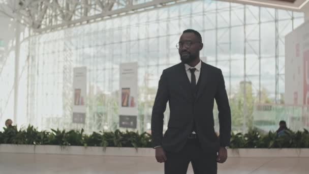 Retrato filmado con seguimiento del empresario afroamericano en ropa formal y gafas posando para la cámara en el centro comercial - Imágenes, Vídeo