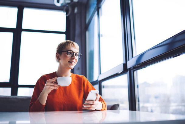 From below of happy female in casual clothes sitting at table with cup of coffee looking out window and typing on smartphone - Фото, изображение