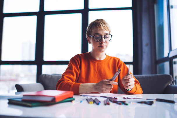 Low angle of focused female artist in casual clothes with eyeglasses sitting at table with notepads choosing colored pencil during work - Photo, image