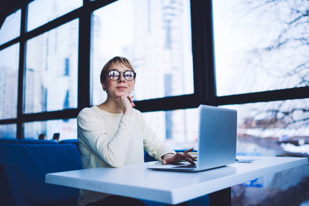 Glad female freelancer in casual clothes with eyeglasses looking at camera sitting at table with hand at chin using laptop while working alone - Fotografie, Obrázek