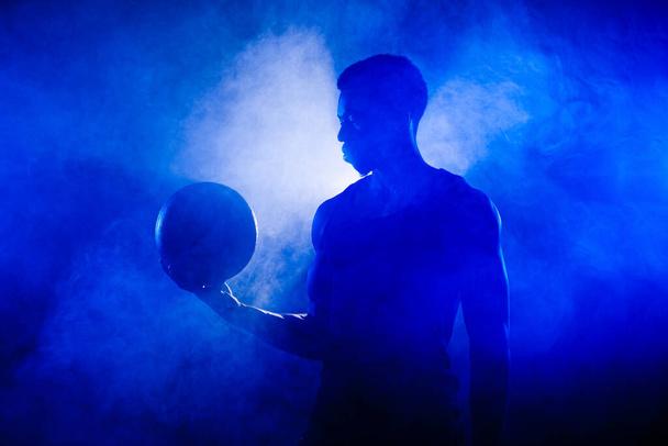 Basketball player holding a ball against blue fog background. African american man silhouette. - Фото, изображение