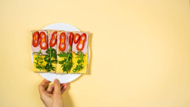 Hand grijpt een witte plaat van vier rechthoekige sandwiches met kaas, ham, paprika en groene arugula van volkoren chips. Gezonde, voedzame gist-vrije snack. Gele achtergrond. - Foto, afbeelding