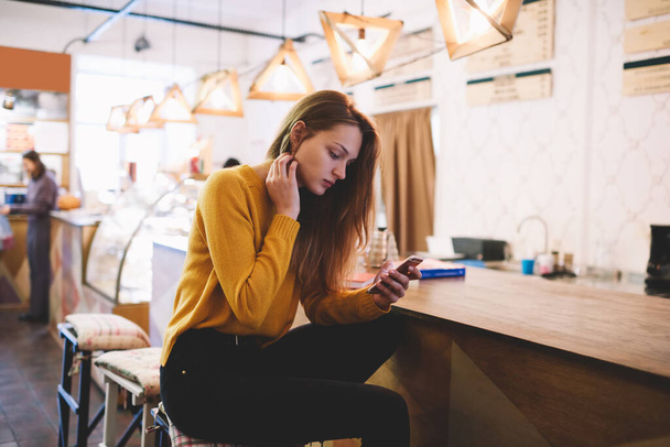 Fiatal gondolkodó nő alkalmi ruhák hosszú haj ül asztalnál szörfözés mobiltelefon töltés szabadidő a modern kávézóban - Fotó, kép