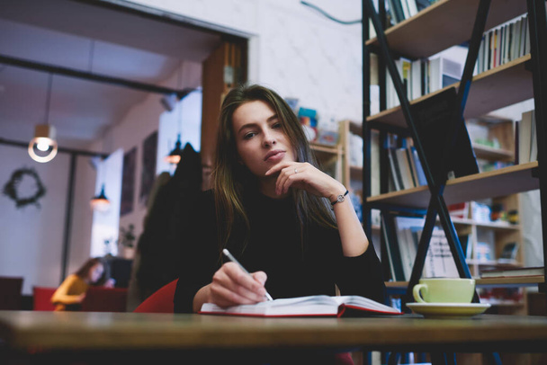 Low angle of focused female student in casual clothes looking at camera touching chin sitting at table with notebook doing homework in library - Fotoğraf, Görsel