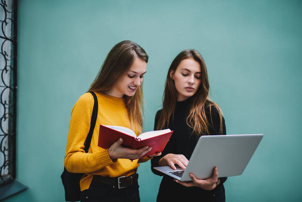 Young female students in casual clothes standing near turquoise wall with opened notepad typing on laptop while doing homework together - Фото, зображення