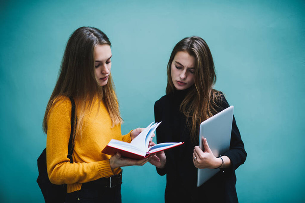 Young pensive female students in casual clothes standing near turquoise wall with modern laptop and notebook discussing home project together - Foto, immagini