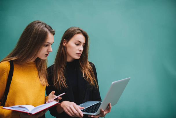 Young thoughtful female students in casual outfits with long hair using smartphone and browsing modern laptop finding information while doing project together - Фото, зображення