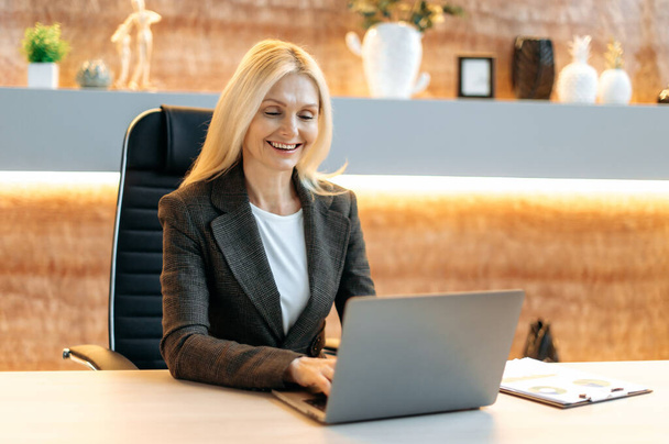 Mature cute happy successful caucasian businesswoman, top manager, company broker, in formal suit, sitting at table in office, using laptop, texting with client or colleagues, smiling friendly - Photo, Image