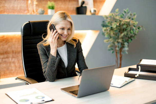 Занятая дружелюбная успешная кавказская бизнесвумен, фрилансер или топ-менеджер, сидящий за столом в офисе с ноутбуком, разговаривающий с клиентом или сотрудниками на мобильном телефоне, улыбающийся - Фото, изображение