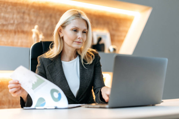 Caucasian mature influential successful smart business woman, financial analyst, manager, sitting at a laptop, studying documents, analyzing diagrams and planning financial strategy - Photo, image