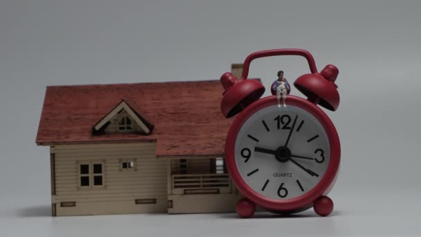 Miniaturní žena sedící s dítětem v červených budících hodinách a dřevěném miniaturním domě. Koncept o ženách, péči o děti a nedostatku času. - Záběry, video