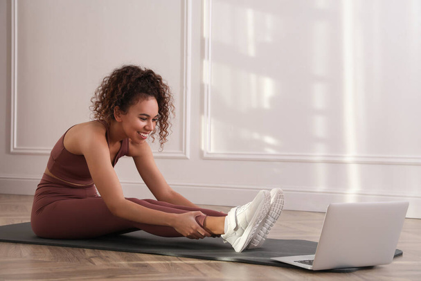 Beautiful African American woman stretching on yoga mat while watching online class indoors - Фото, изображение
