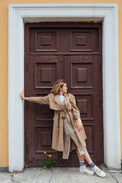 Young smiling millennial woman with wild hair dressed in an autumn coat posing near the door of an old building. - Zdjęcie, obraz