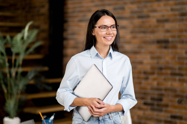 Portrait beautiful smart confident young adult brunette caucasian woman, office manager, teacher, broker wearing glasses, standing in office, holding laptop in her hands, looking to side, smiling - Foto, Imagem