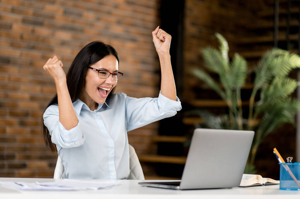Happy joyful successful smart emotional caucasian business woman, broker, freelancer wearing glasses, sitting in office with laptop enjoying good deal, victory, good news, gesturing with fists smiling - Photo, Image