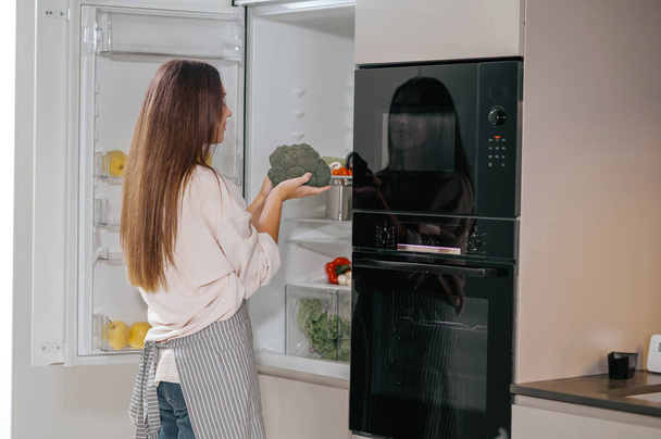 Jonge huisvrouw staat bij de koelkast in de keuken - Foto, afbeelding