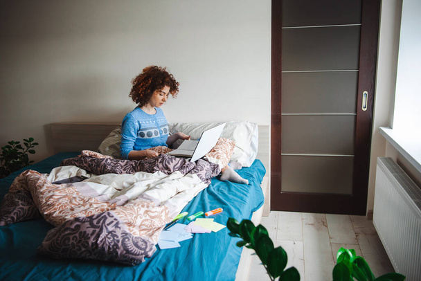 Mulher encaracolada caucasiana fazendo aulas on-line da cama usando laptop em um pijama azul. Estilo de vida de quarentena. - Foto, Imagem