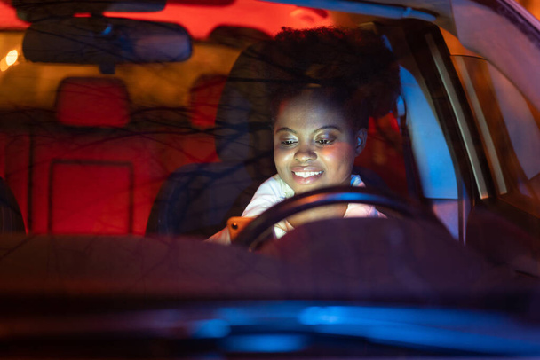 Woman typing in smartphone driving car. Black girl read sms on phone sitting on driver seat at night - Fotó, kép