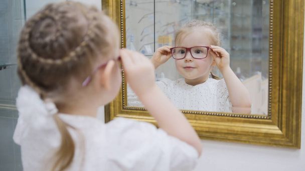 Klein meisje probeert nieuwe glazen in de buurt van spiegel - winkelen in oogheelkunde kliniek - Foto, afbeelding