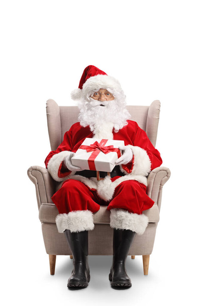 Full length portrait of santa claus sitting in an armchair with a present in his hands isolated on white background - Photo, Image