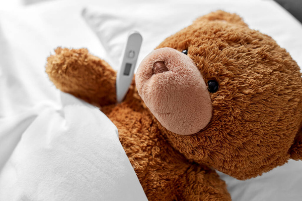 ill teddy bear toy head with thermometer in bed - Foto, afbeelding